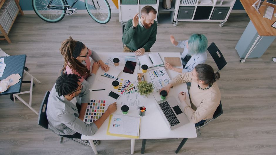 Vista superior de uma equipe criativa diversificada colaborando em um espaço de trabalho com tecnologia e gráficos.